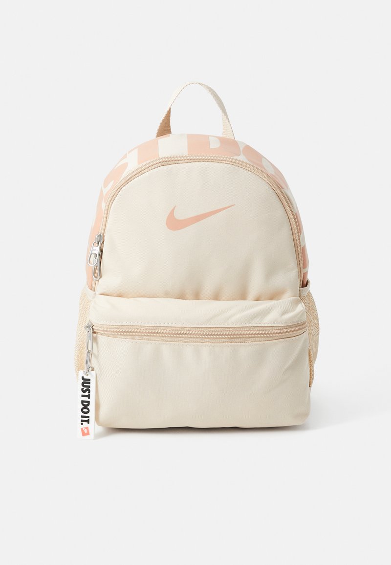 Beige backpack with light pink accents, featuring a prominent Nike logo, zippered front pocket, and woven "JUST DO IT" tag. Textured fabric.