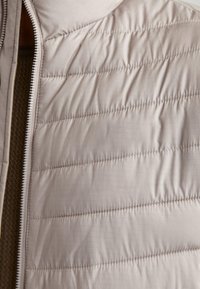 Light beige puffer vest featuring a quilted texture, zippered front, and a smooth satin-like finish, with subtle reflective accents.