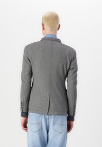 Grauer Blazer mit strukturierter Oberfläche, ausgestattet mit einem Reverskragen und einem einzelnen Schlitz. Kombiniert mit hellblauen Denim-Jeans. Knopf-Details an den Ärmeln.