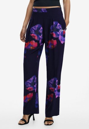 Paarse bloemenbroek met grote roze en blauwe bloemenpatronen, plooien aan de voorkant en zijzakken. Gecombineerd met zwarte sandalen met bandjes.