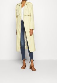 Žlutý trenčkot s dvojitým zapínáním, dlouhými rukávy a volným střih, zkombinovaný s modrými skinny džíny a hnědými sandály na páse.