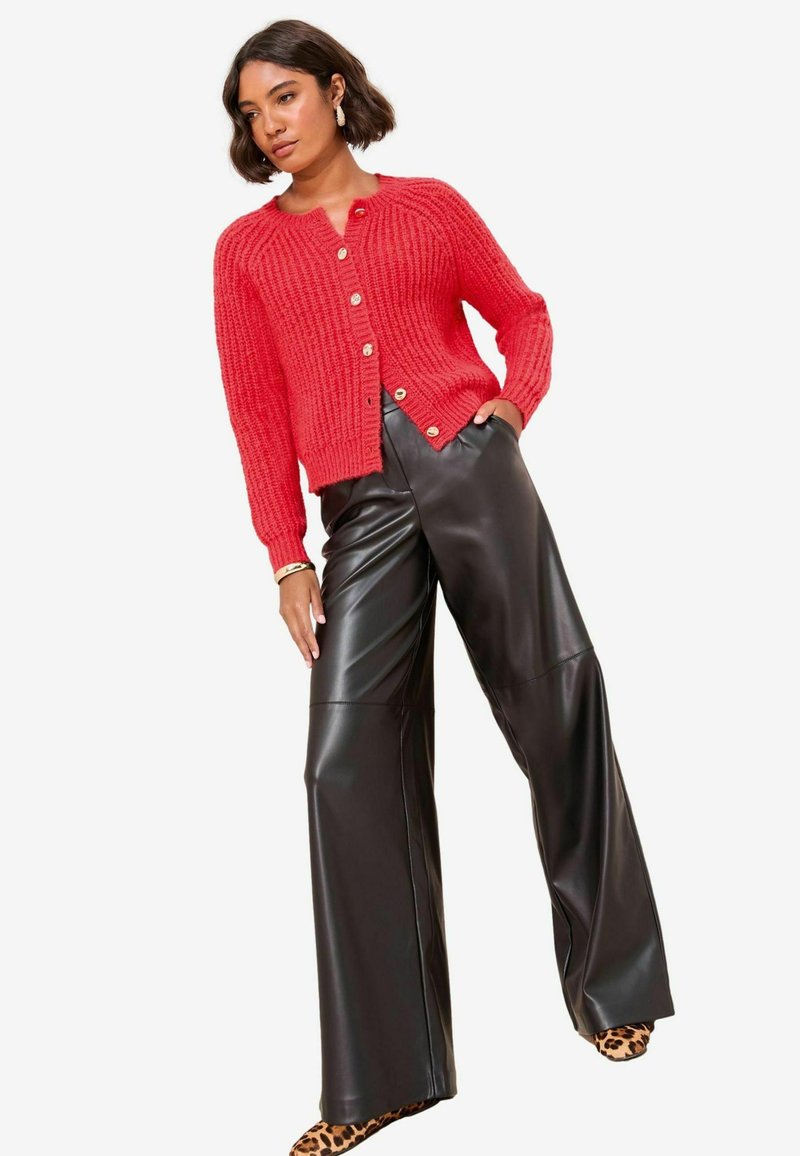 Cardigan rosso lavorato a maglia con texture a coste e bottoni dorati, abbinato a pantaloni neri in ecopelle a vita alta e scarpe con stampa leopardata.