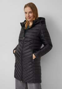 Zwarte pufferjas met ritssluiting, capuchon en een chevron-quilting patroon. Beschikt over twee zijzakken en een middellange snit.