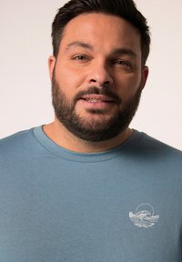 Camiseta de algodón en color azul verdoso con cuello redondo y mangas cortas. Presenta un gráfico de olas blancas en la zona izquierda del pecho. Textura suave.