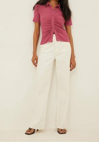 Różowa koszula z krótkim rękawem, zapinana na guziki, z marszczeniem na środku, w połączeniu z szerokimi białymi spodniami i czarnymi sandałami na obcasie.