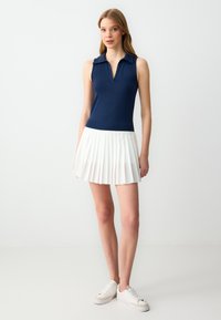 Polo senza maniche navy con scollo a V, abbinato a una gonna plissée bianca. L'outfit è completato da sneakers bianche.