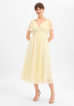 Frau, die ein blassgelbes, knielanges Kleid mit transparenten, flatternden Ärmeln und goldenen Sandalen mit hohen Absätzen trägt, vor weißem Hintergrund.