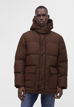 Homme portant une doudoune marron avec de grandes poches avant et un col montant, associée à un jean bleu sur un fond uni.