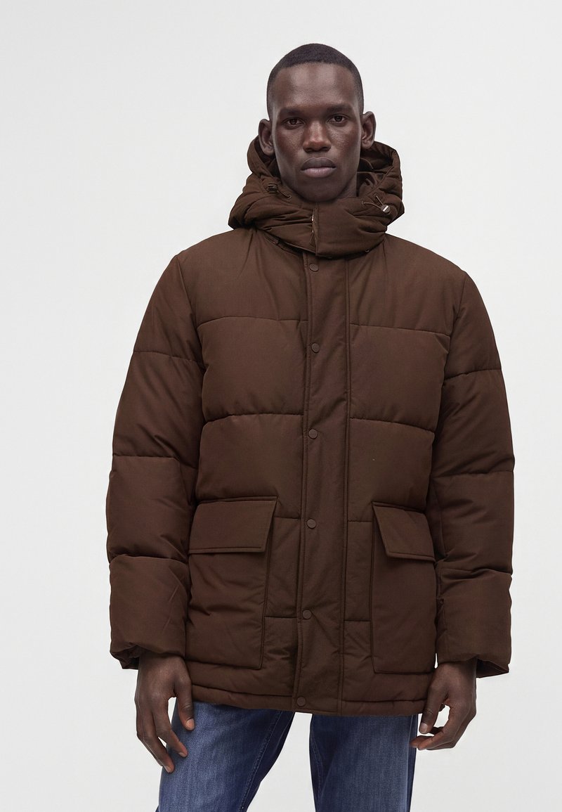 Homme portant une doudoune marron avec de grandes poches avant et un col montant, associée à un jean bleu sur un fond uni.