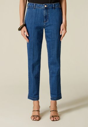 Frau trägt gerade geschnittene blaue Jeans, schwarzes Top, braunes Armband und offene High-Heel-Sandalen mit Schlangenmuster, steht vor beigem Hintergrund.