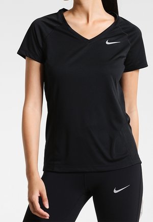 Kobieta ubrana w czarną sportową koszulkę z krótkim rękawem i dekoltem w serek oraz czarne legginsy, oba z białymi logo Nike na klatce piersiowej i udzie.
