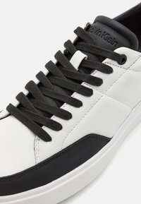 Sneakers en cuir blanc avec une semelle en caoutchouc noir. Lacets et accents noirs sur la languette et le côté. Surface texturée et détails de couture notables.