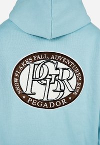 Ανοιχτό μπλε φούτερ με κουκούλα, διακοσμημένο με έναν μεγάλο οβάλ κεντητό λεκέ που φέρει τις λέξεις "ΠΤΩΣΕΙΣ ΧΙΟΝΙΟΥ, ΑΝΑΒΑΣΕΙΣ ΠΕΡΙΠΛΑΝΗΣΕΩΝ" και "PEGADOR" σε καφέ και λευκό.