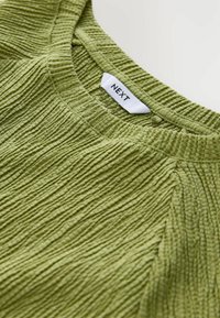 Grön texturerad topp med vertikal ribbstruktur, med rund halsringning och etiketten "NEXT" sydd i vitt. Mjuk tyg med lätt stretch.