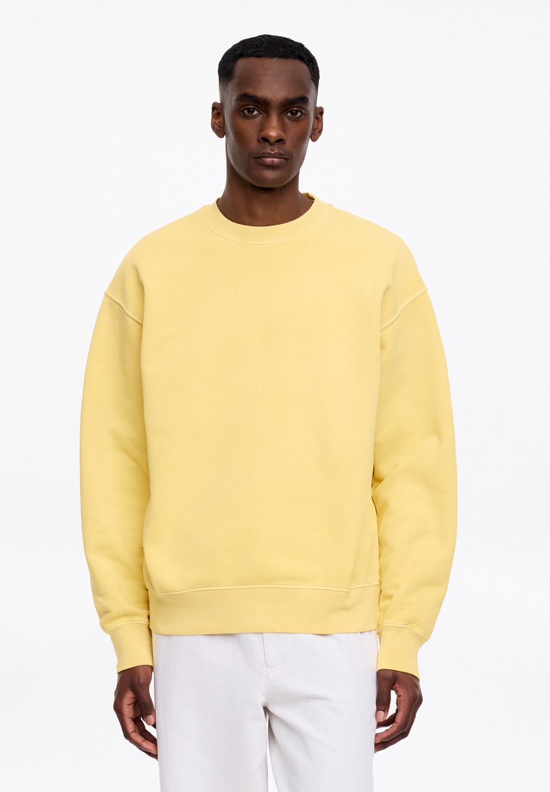 Mann trägt ein schlichtes gelbes Sweatshirt mit Rundhalsausschnitt und weiße Hose, steht vor weißem Hintergrund.