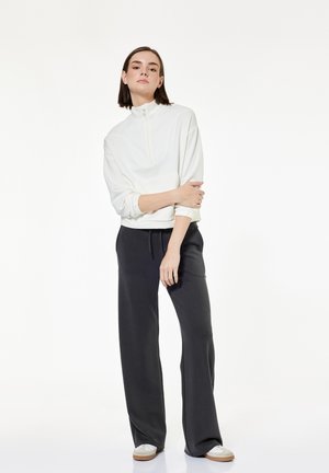 Kobieta stoi z założonymi rękami, ubrana w biały pulower z ćwierćzamkiem, luźne czarne spodnie i białe trampki na jednolitym tle.