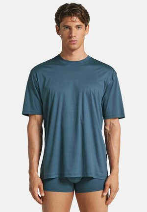 Ung mand med kort brunt hår iført en løs blå t-shirt med rund hals og matchende blå boxershorts mod en ensfarvet baggrund.