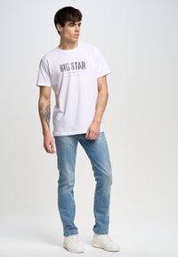 Camiseta de algodón blanca con gráfico de "BIG STAR" en impresión oscura. Vaqueros de mezclilla azul claro y zapatillas blancas, de pie contra un fondo sencillo.