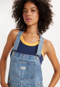 Τζιν σαλοπέτα με ρυθμιζόμενες τιράντες, σε ανοιχτό μπλε χρώμα, με μπροστινή τσέπη και ετικέτα. Φορεμένη πάνω από μια ναυτική μπλε και κίτρινη ριμπ μπλούζα.