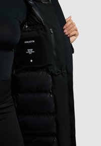 Zwarte gewatteerde vest met een matte stof en glanzende accenten, voorzien van een ritszak en een merkembleem aan de zijkant.