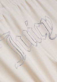 Krēmkrāsas audums ar viļņainu rakstu, ko veido sudraba rhinestones. Tekstūra ir gluda, ar savērunāmiem detaļām augšpusē.