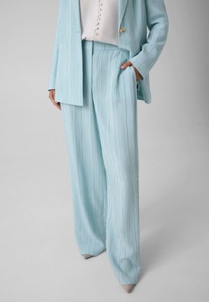 Persona que lleva pantalones anchos de rayas finas en azul claro y blazer a juego sobre camiseta blanca, de pie con una mano en el bolsillo.