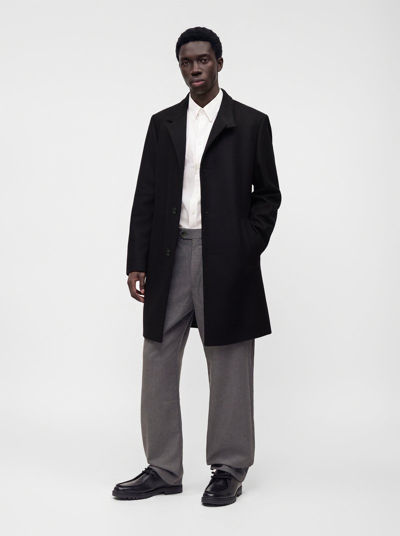 Jeune homme portant un manteau noir, une chemise blanche, un pantalon gris et des chaussures noires, debout devant un fond clair uni.