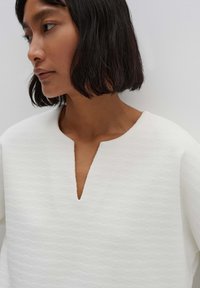 Blusa bianca texturizzata con scollatura a V e vestibilità ampia. Presenta sottili strisce orizzontali e maniche larghe. Aspetto del tessuto liscio.