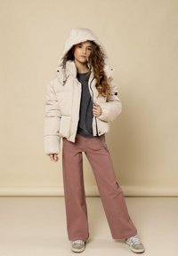 Beige pufferjas met een capuchon, voorzien van grote doorgestikte delen en een ritssluiting aan de voorkant. Gecombineerd met roze wijde broek en grijze sneakers.