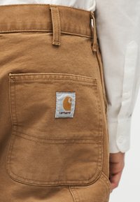 Πίσω τσέπη καφέ παντελονιών Carhartt που φοριούνται με λευκό πουκάμισο μακρύ μανίκι, δείχνοντας την υφή του υφάσματος και τις ραμμένες ραφές.