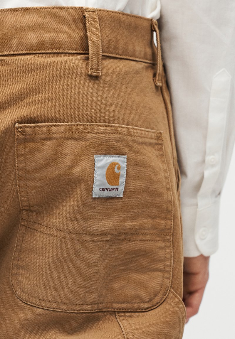 Poche arrière d'un pantalon Carhartt marron porté avec une chemise à manches longues blanche, montrant la texture du tissu et les coutures cousues.