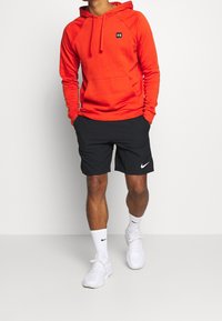 Felpa rossa con cappuccio e tasca frontale e logo nero, abbinata a pantaloni corti neri con un baffo bianco Nike, indossata con scarpe bianche.