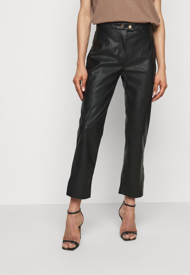 Femme portant un pantalon court taille haute en simili cuir noir, accompagné de sandales noires à talons hauts et d'un haut marron.