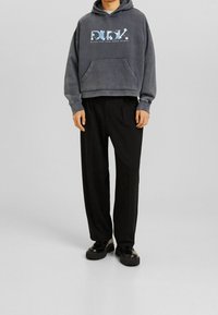 Felpa grigia con cappuccio, tasca frontale e testo grafico, abbinata a pantaloni neri plissettati e scarpe nere chunky. Tessuto liscio, vestibilità comoda.