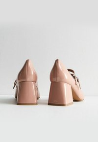 Jasnoróżowe lakierowane buty na słupku z ozdobną kokardą i srebrnymi detalami, gładka faktura i zaokrąglony fason palców.