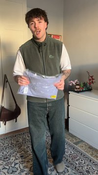 Grijze fleece bodywarmer over een wit t-shirt, een lichtpaarse shirt met print in verpakking vasthoudend. Blauw jeans met relaxte pasvorm en beige schoenen.