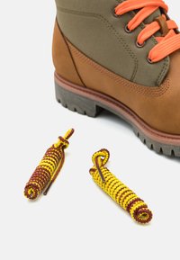 Botte en cuir marron et toile olive avec des lacets orange. Deux embouts de lacet en jaune et bordeaux enroulés sont placés sous la botte.