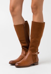 Bottes en cuir et daim marron jusqu'au genou avec une fermeture éclair latérale et un détail de bride à la cheville. Talon plat, bout arrondi, texture lisse.