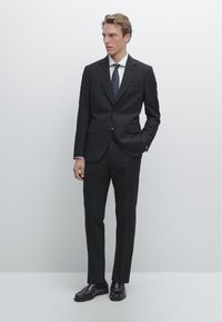 Dunkelgrauer maßgeschneiderter Anzug mit einem einstufigen Blazer, passenden Hosen und einem weißen Hemd. Schwarze Lederschuhe vervollständigen den Look.