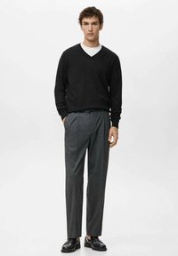 Man draagt een zwarte V-hals trui over een wit overhemd, grijze geplooide broek en zwarte leren loafers, staand met één hand in de zak.