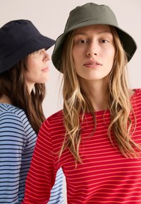 Zwei Frauen tragen Bucket Hats; einer ist dunkelblau, der andere olivgrün. Sie tragen gestreifte Oberteile; rot-orange gestreift, blau-schwarz gestreift.