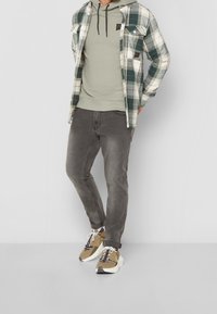 Camisa de cuadros en blanco y verde, superpuesta sobre una sudadera verde oliva, combinada con jeans grises y zapatillas beige con acentos oscuros.