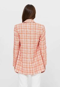 Donna con lunghi capelli castani lisci che indossa un blazer aderente a quadretti arancioni e bianchi, rivolta di spalle su uno sfondo semplice.