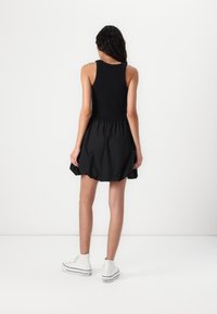Zwarte mouwloze jurk met een getailleerde top en een uitlopende rok, gemaakt van lichte stof. G gedragen met witte hoge sneakers.