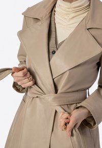 Béžový trench coat z umělé kůže s širokým límcem, opaskem v pase a dlouhými rukávy, vrstvený přes kostkovanou vestu a světlý top.