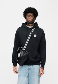 Μαύρο φούτερ με μακριά μανίκια και μπροστινή τσέπη, που διαθέτει κυκλικό λογότυπο. Συνδυασμένο με ανοιχτό μπλε τζιν και μαύρη τσάντα με λευκές λεπτομέρειες.