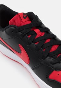 Black synthetic sneaker with red mesh accents and laces, featuring a prominent red Nike swoosh and white outsole. Textured surfaces.