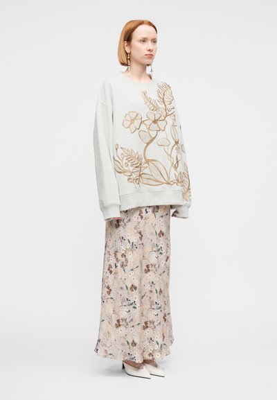 Γυναίκα με κοντά κόκκινα μαλλιά που φοράει φαρδιά μπεζ μπλούζα με καφέ ανθιστή κεντήματα, μακριά φλοράλ φούστα, λευκά τακούνια και κρεμαστά σκουλαρίκια.