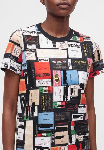 Άτομο που φοράει πολύχρωμο t-shirt Moschino καλυμμένο με διάφορα τυπωμένα σχέδια υφασμάτινων ετικετών σε πολλαπλά χρώματα και γραμματοσειρές.