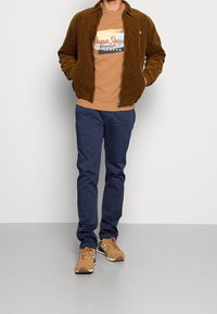 Un uomo con giacca in velluto a coste marrone, felpa grafica beige, pantaloni blu navy e sneakers beige in piedi contro un muro bianco.